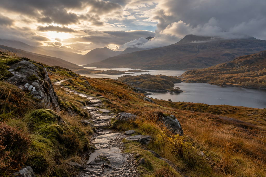 Herbstliche Landschaft in den schottischen Highlands mit einem steinigen Wanderweg, goldbraunen Gräsern, dunklen Lochs und nebelverhangenen Bergen unter dramatischem Wolkenhimmel.